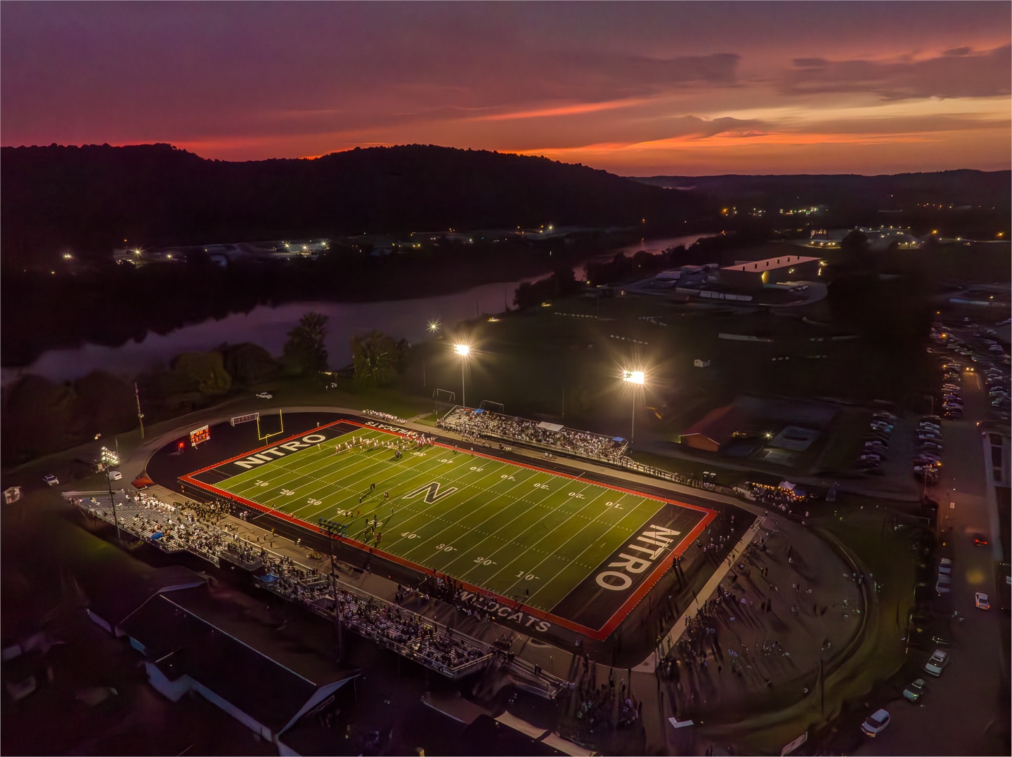 Aerial view of a high school football game at sunset, with stadium lights illuminating the field.