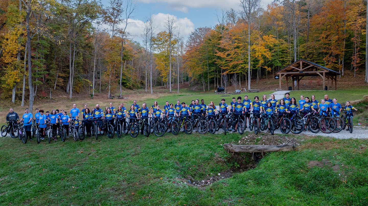 Group of mountain bikers in blue and yellow jerseys posing with their bikes in a forest setting during autumn.