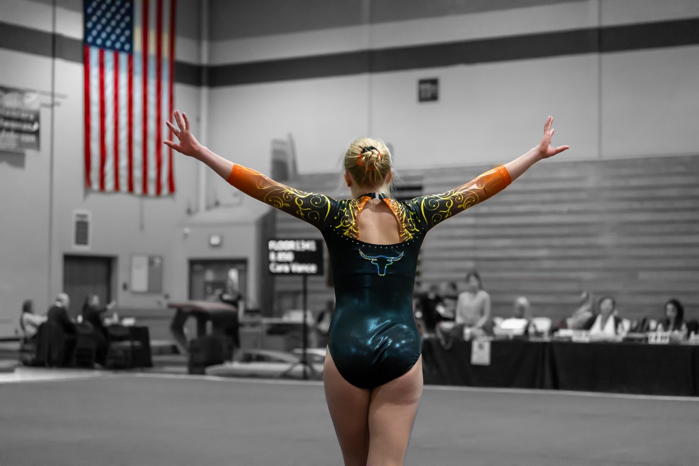 Female gymnast finishing her routine with a confident pose in front of the American flag at a gymnastics competition.