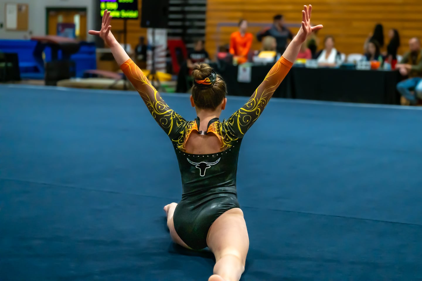 Gymnast in a black and orange leotard with arms raised in a seated pose on a gymnastics floor.