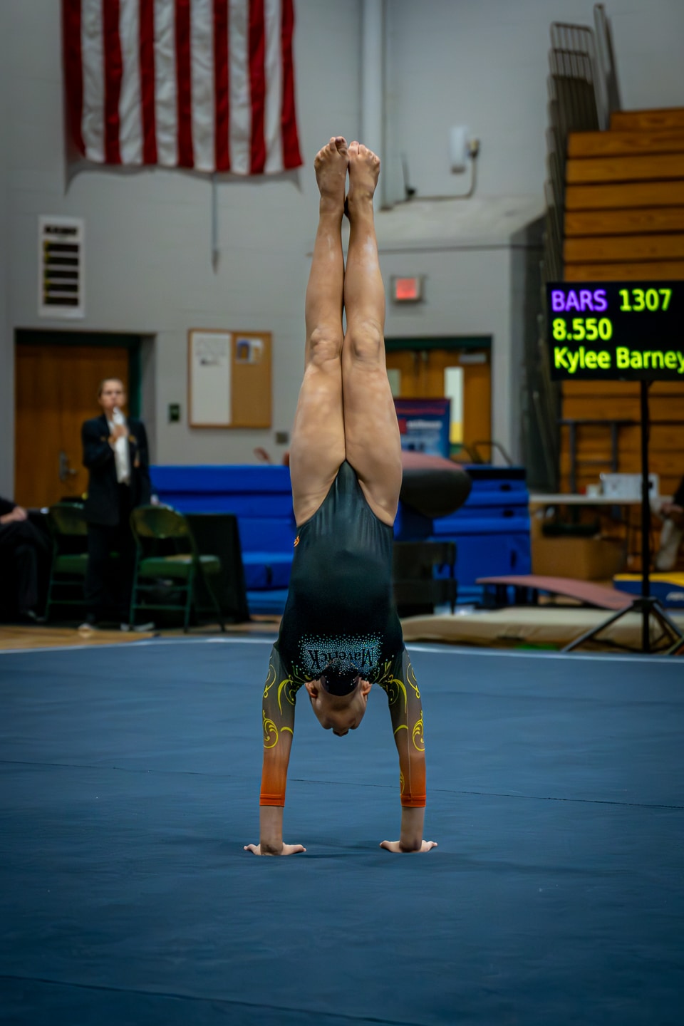 Gymnast performing a handstand at a gymnastics event, with the American flag in the background.