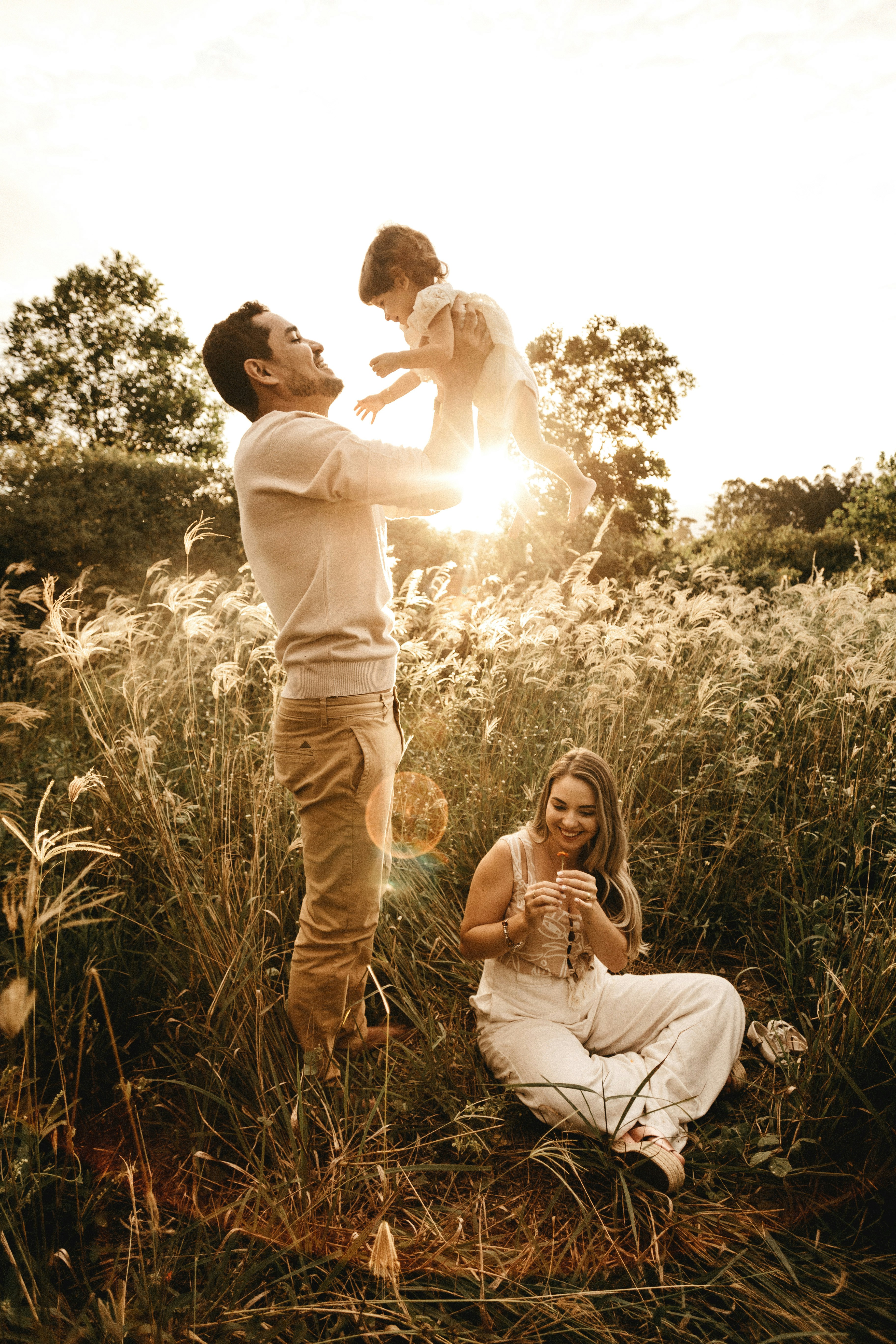 Family enjoying a sunset field photoshoot with father lifting child high and mother smiling on the ground.