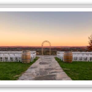 Framed-Elegant Outdoor Wedding Venue in Autumn | A stone pathway flanked by rows of white chairs and wine barrels leads to a romantic wedding arch overlooking a scenic landscape with an autumn sunset.