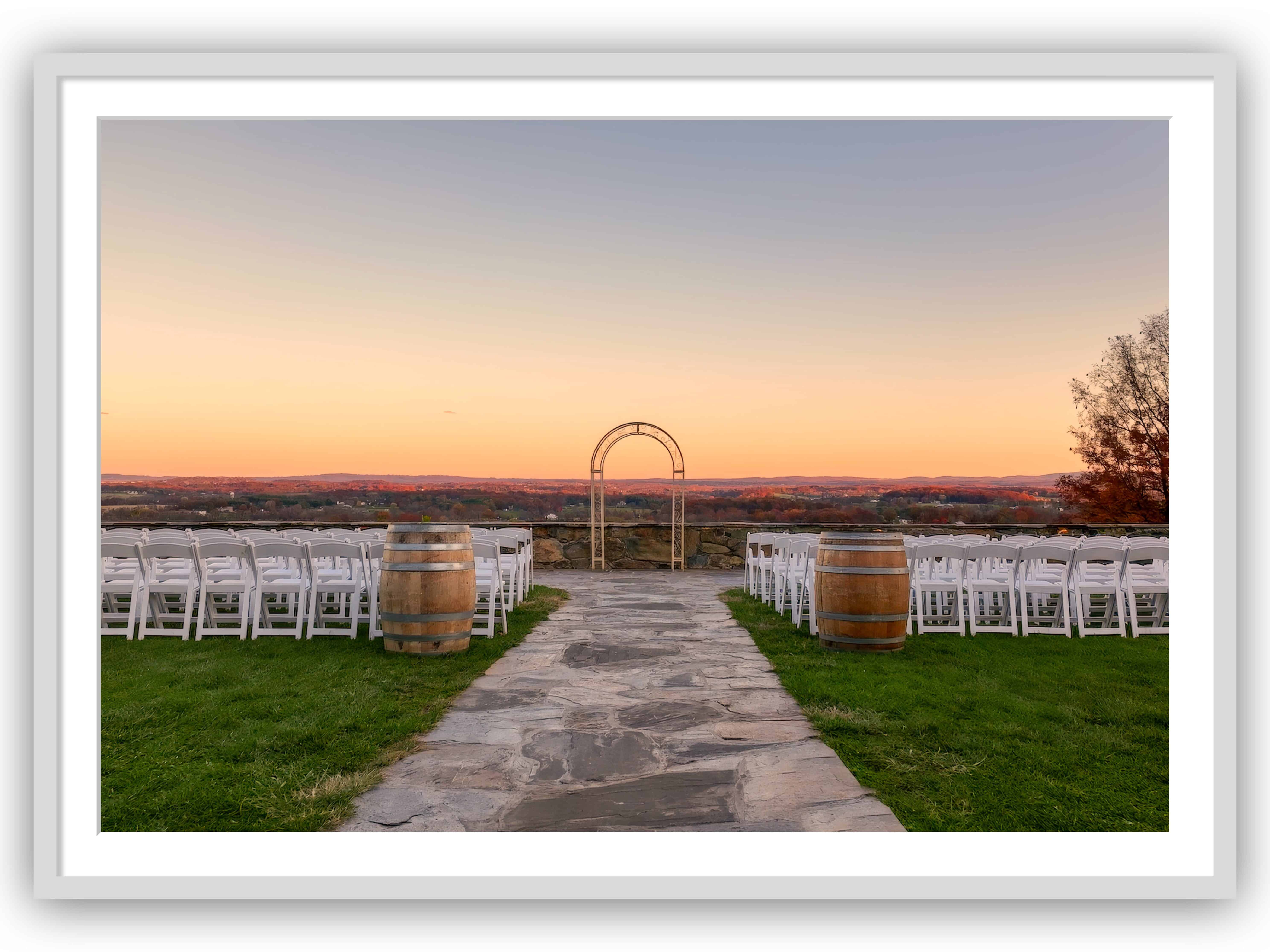 Framed-Elegant Outdoor Wedding Venue in Autumn | A stone pathway flanked by rows of white chairs and wine barrels leads to a romantic wedding arch overlooking a scenic landscape with an autumn sunset.